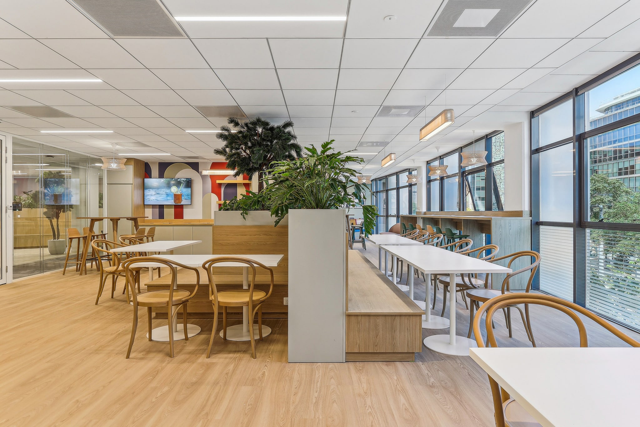 Cafétéria de bureau design avec mobilier en bois, tables blanches et plantes vertes sous de grandes baies vitrées.