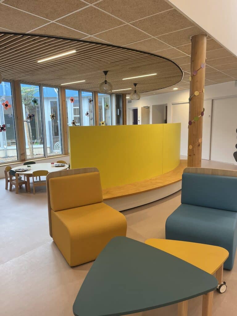 Aménagement intérieur d'une crèche moderne avec mobilier coloré, mur jaune courbé et coin repos lumineux.