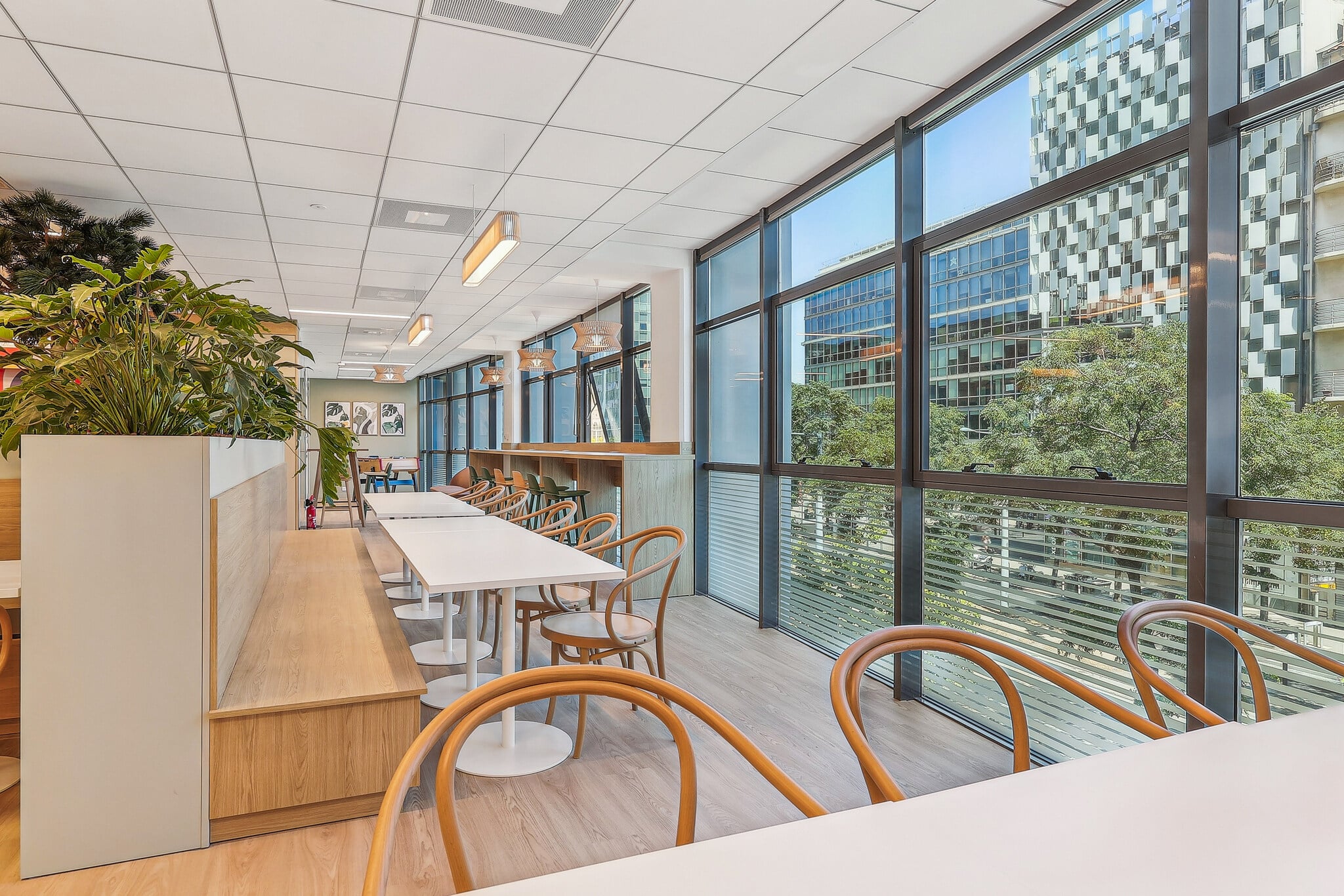 Espace café moderne avec grandes baies vitrées, tables blanches et chaises en bois dans un bureau lumineux.
