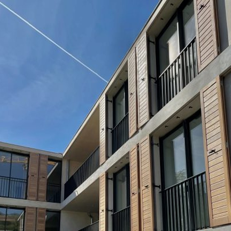 Façade moderne d'un immeuble à deux étages avec balcons noirs, baies vitrées et volets persiennes en bois clair sous un ciel bleu traversé par une traînée d'avion.