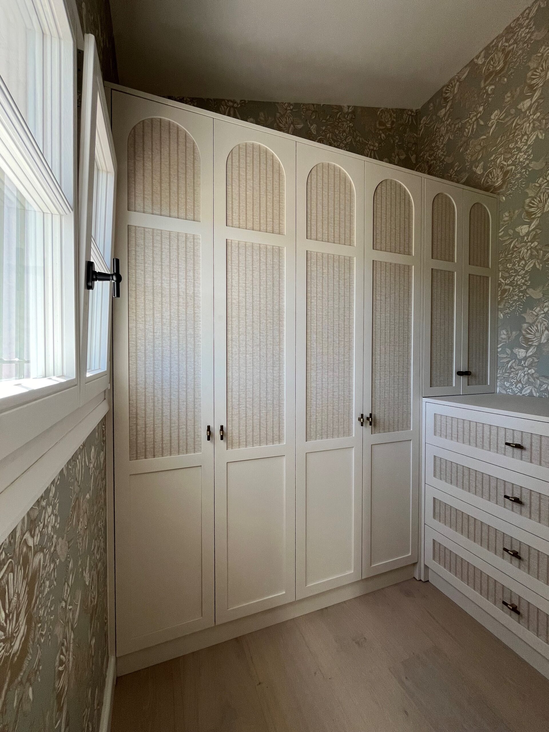 Armoire blanche à arches et panneaux en rotin, commode assortie sur fond de papier peint floral vintage.