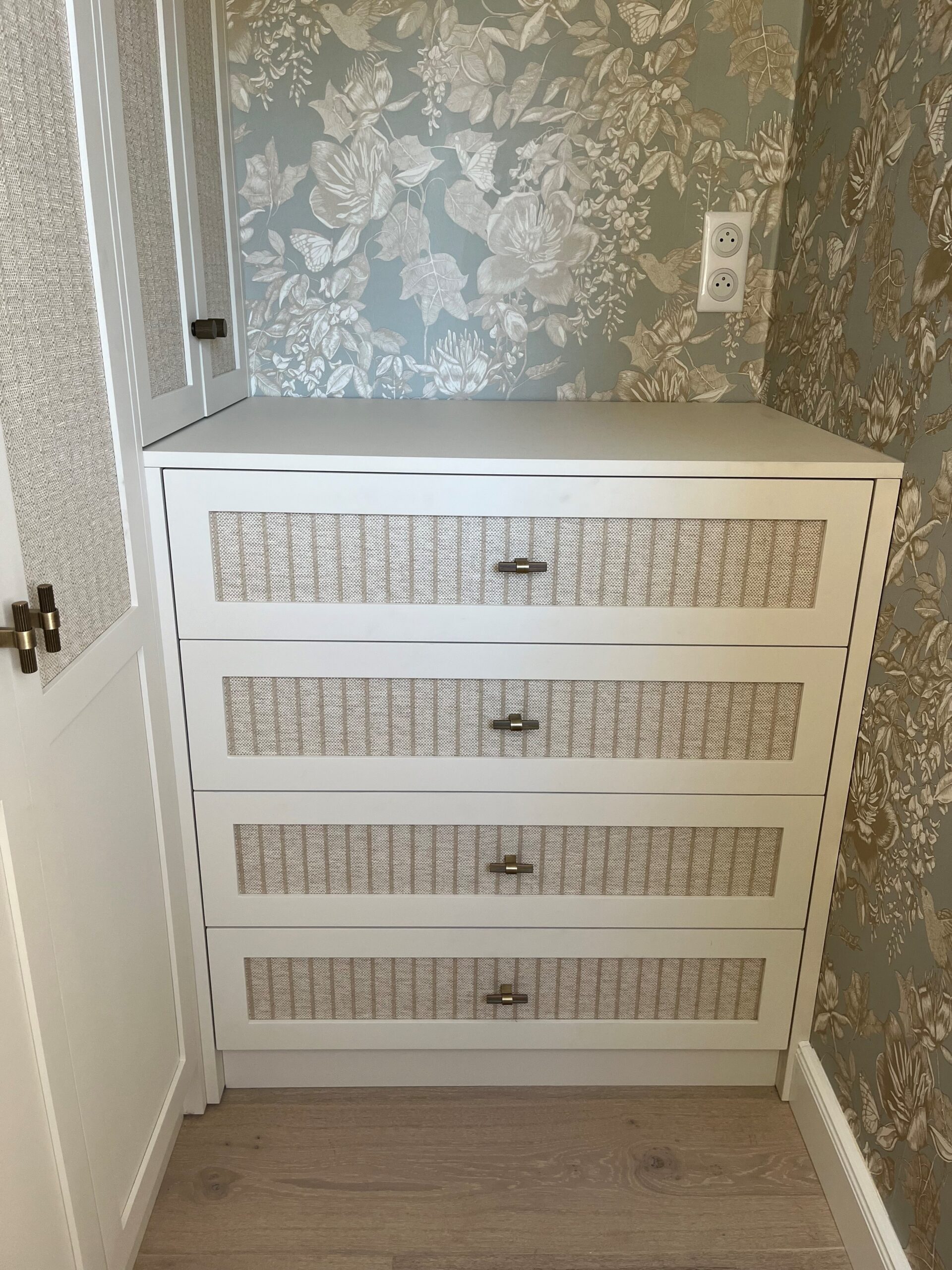Commode blanche à quatre tiroirs avec panneaux en rotin et poignées dorées devant un papier peint floral chic.