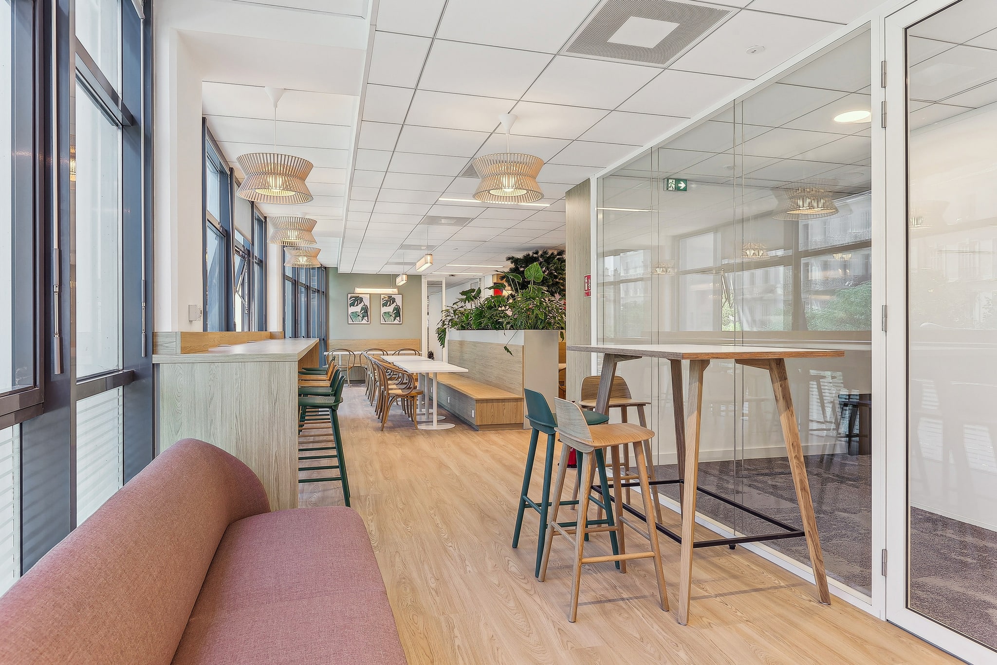Espace de coworking moderne et lumineux avec mobilier en bois, canapé rose, tables hautes et grandes baies vitrées.