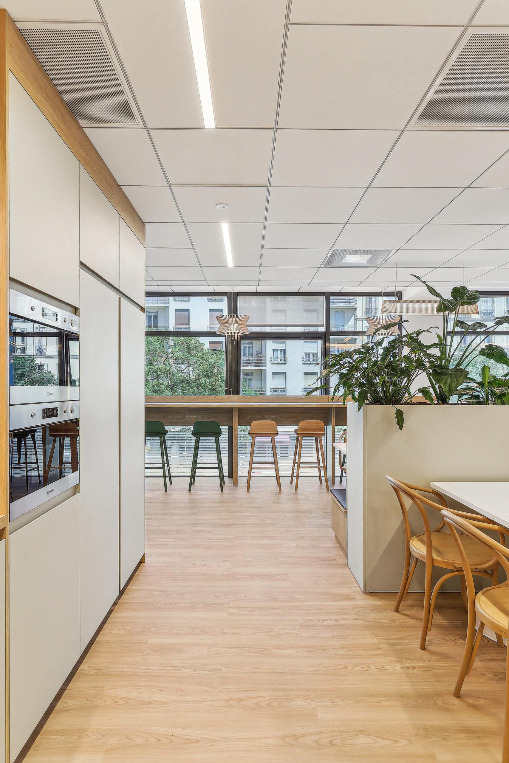 Cuisine de bureau moderne avec meubles blancs, tabourets en bois et bar offrant une vue urbaine lumineuse.