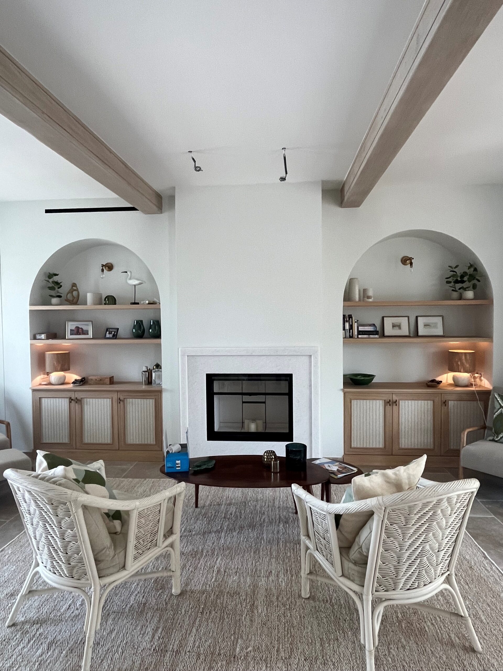 Salon lumineux avec cheminée, niches en arche, étagères en bois et fauteuils en rotin blanc.