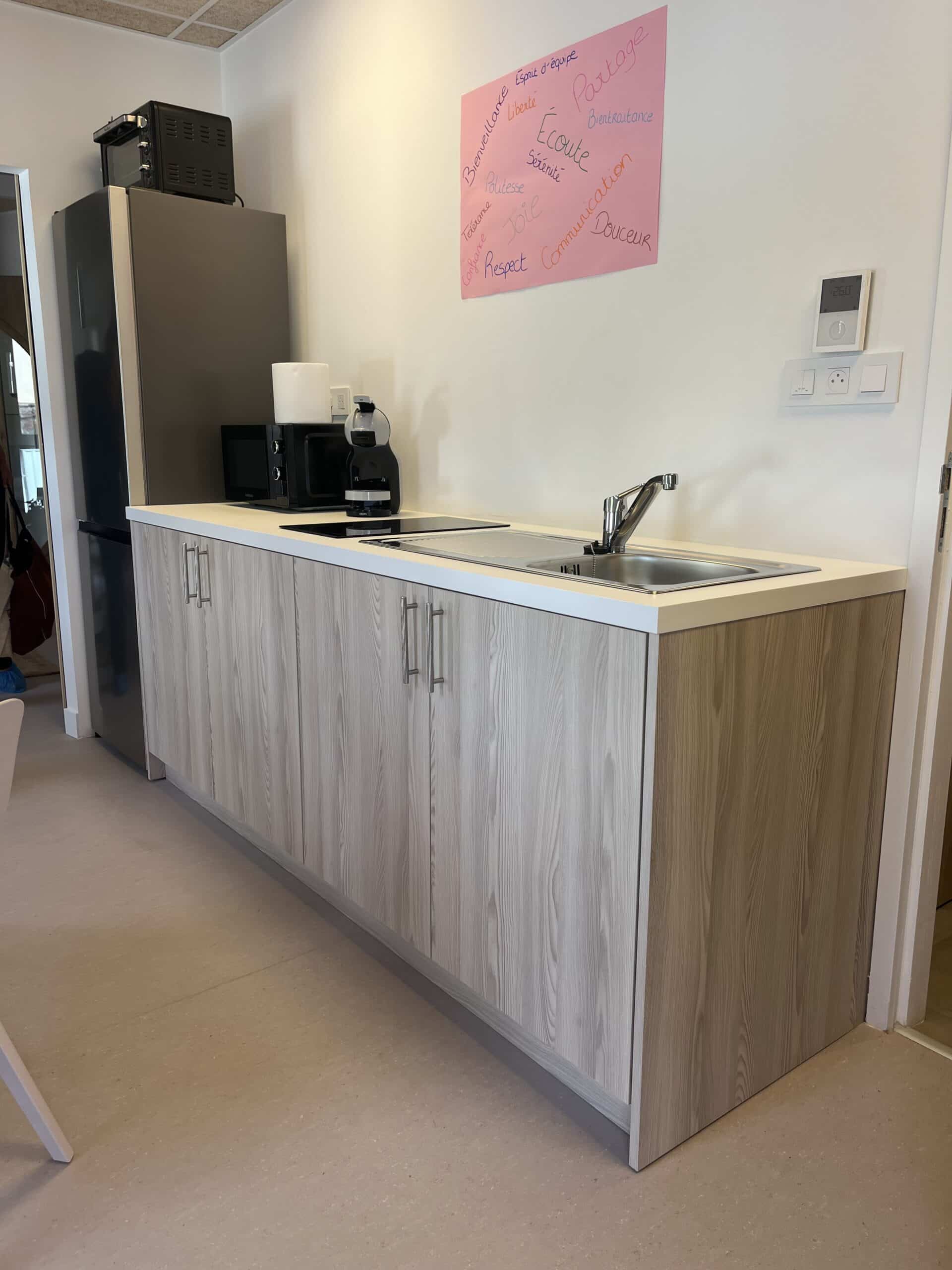 Kitchenette moderne en bois clair avec évier, plaque, électroménager et affiche rose de valeurs d'équipe au mur.