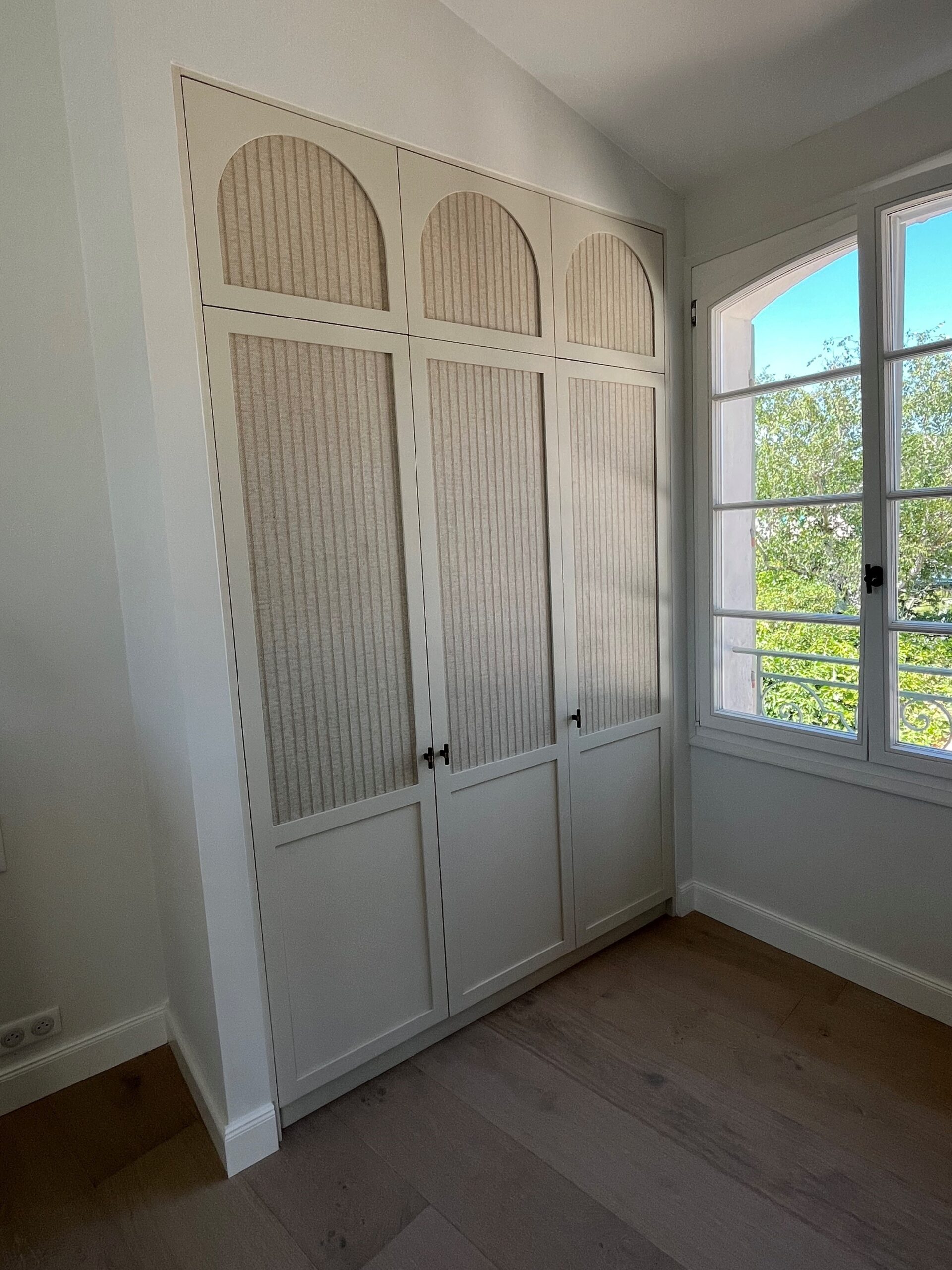Placard sur mesure beige avec arches et texture cannelée dans une chambre lumineuse à côté d'une fenêtre.