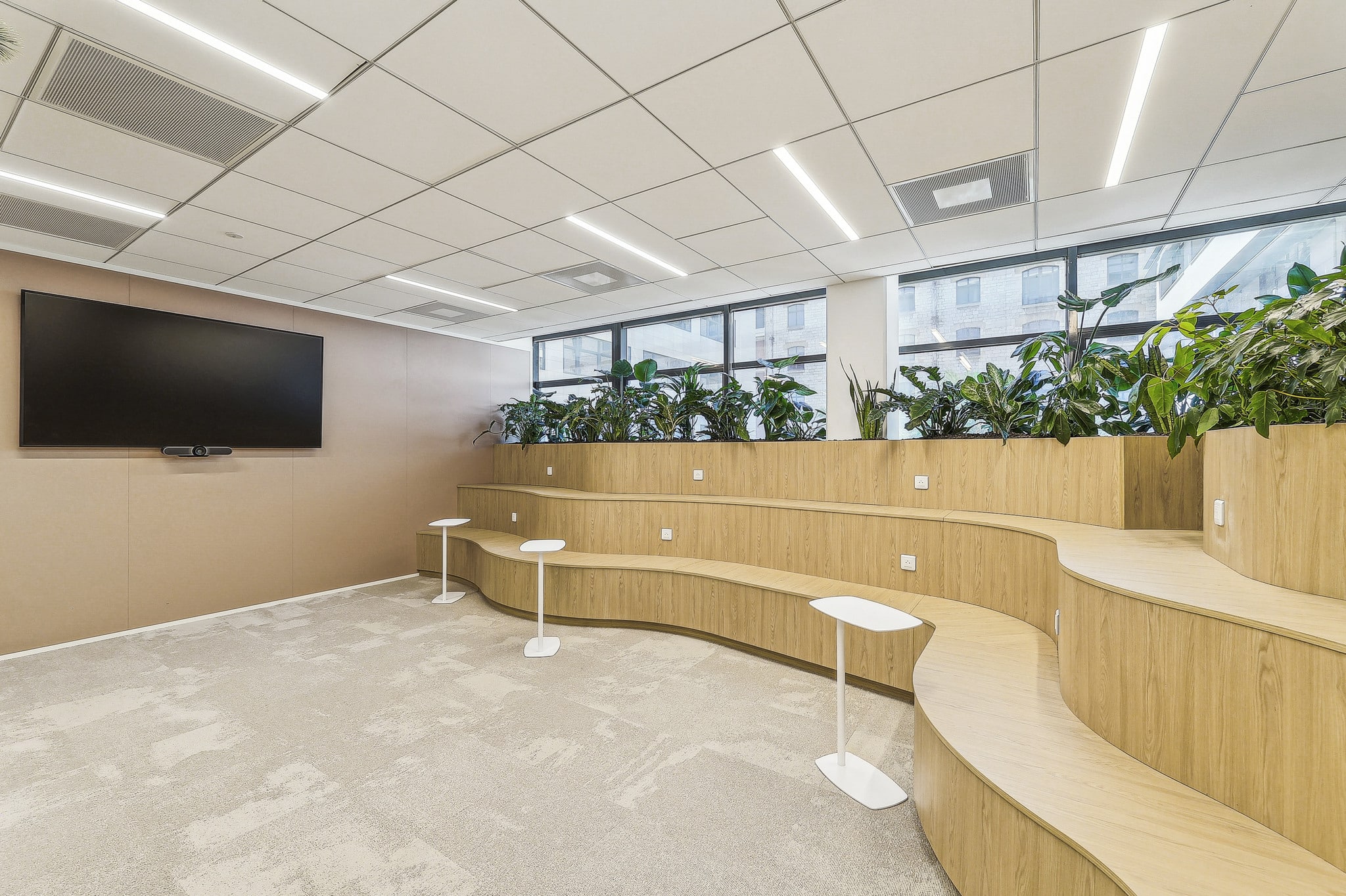Gradins en bois clair dans une salle de réunion moderne avec plantes, écran mural et tables d'appoint blanches.