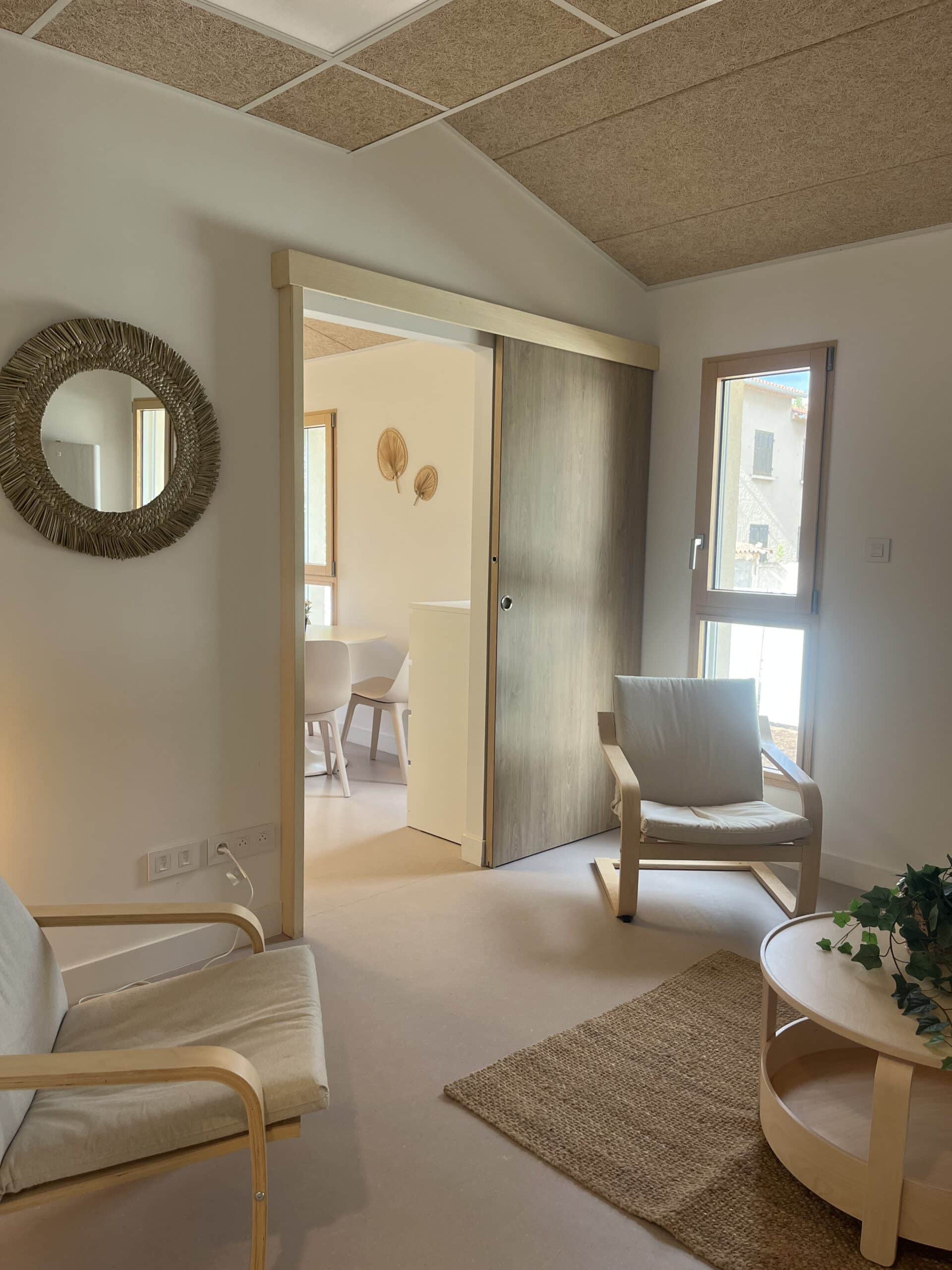 Salon scandinave avec fauteuils bois, miroir osier et porte coulissante, ambiance lumineuse et minimaliste.