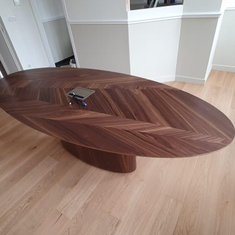Grande table ovale en noyer avec marqueterie chevrons et trappe technique ouverte, reposant sur un pied central, sur parquet clair.