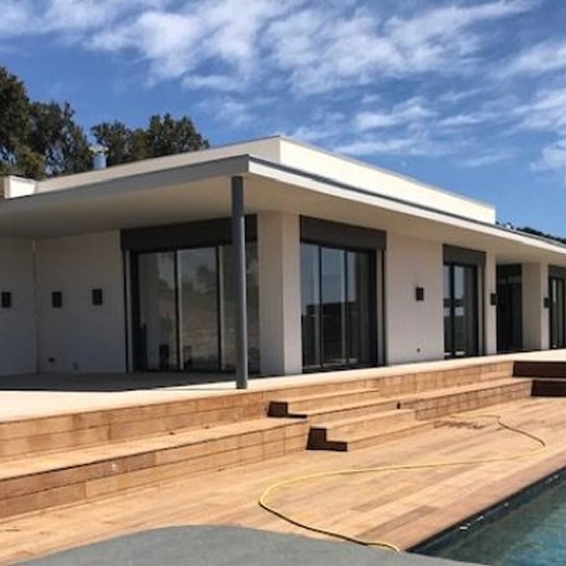 Villa moderne blanche avec toit plat et larges baies vitrées, terrasse en bois à plusieurs niveaux près d'une piscine sous un ciel bleu nuageux.