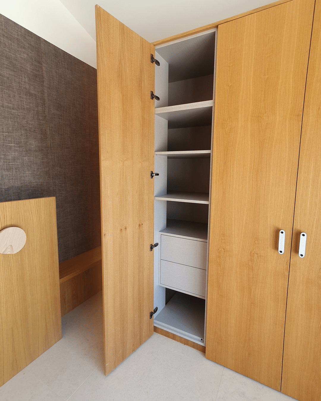 Gros plan sur une armoire en bois clair ouverte, révélant des étagères et deux tiroirs gris, près d'un mur texturé marron.