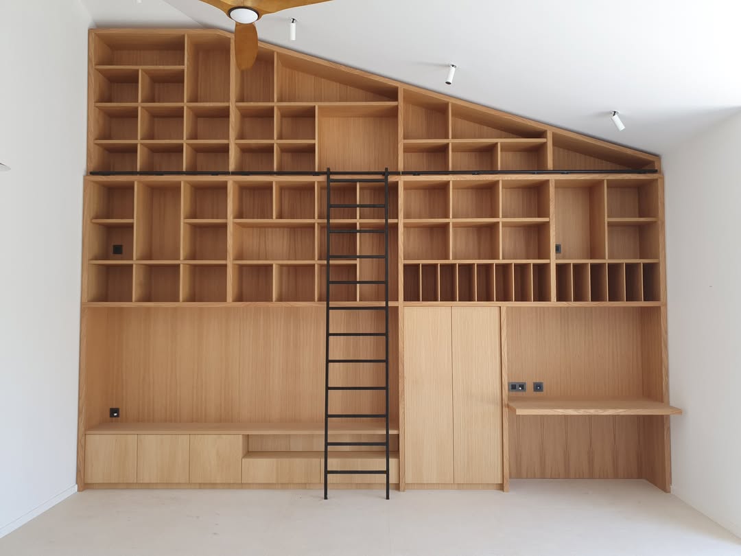 Grande bibliothèque intégrée en bois clair, avec étagères inclinées, échelle noire coulissante, coin bureau et rangements bas.