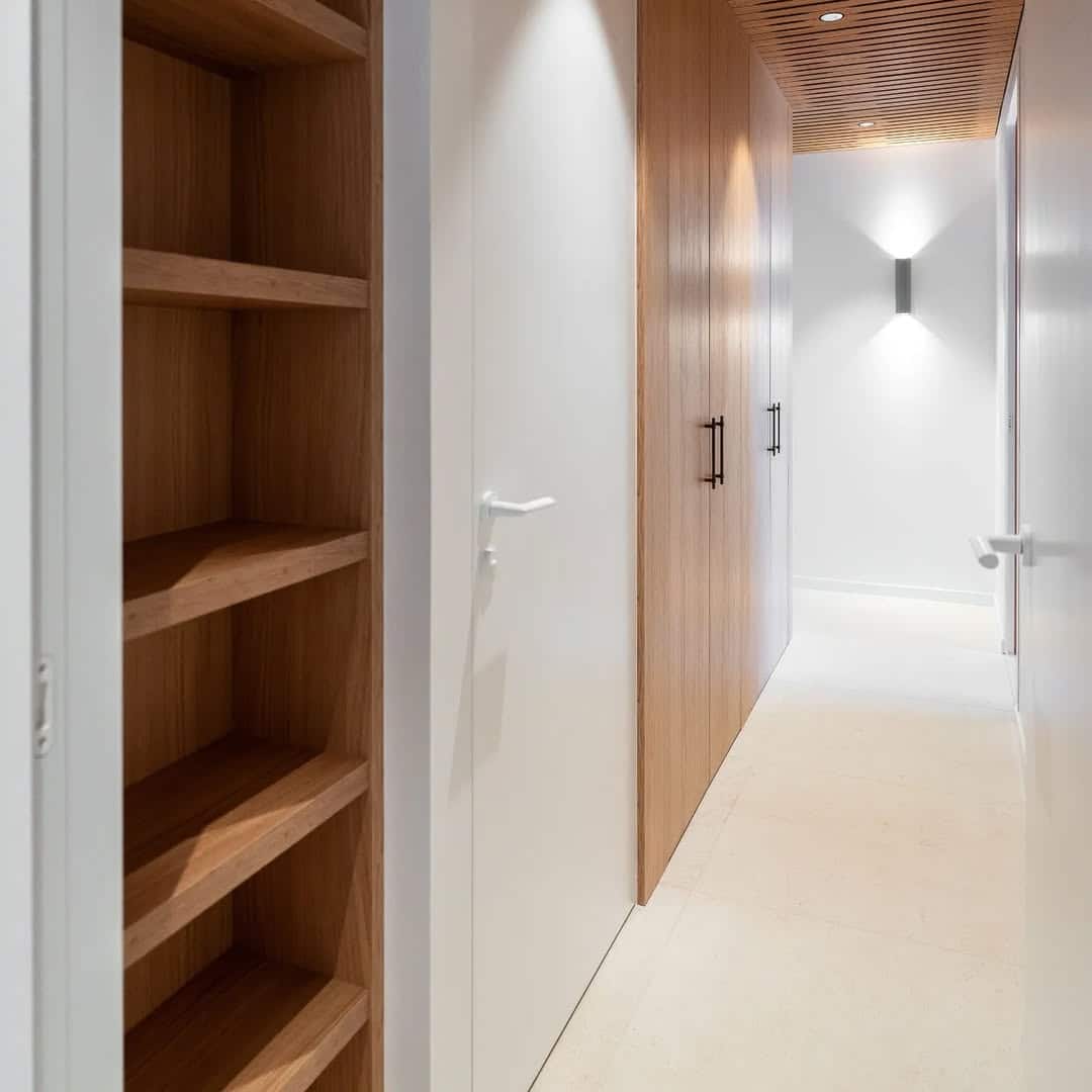 Couloir moderne avec étagères en bois clair, portes blanches à poignées blanches, et un placard en bois avec poignées noires. Plafond à lattes boisées.
