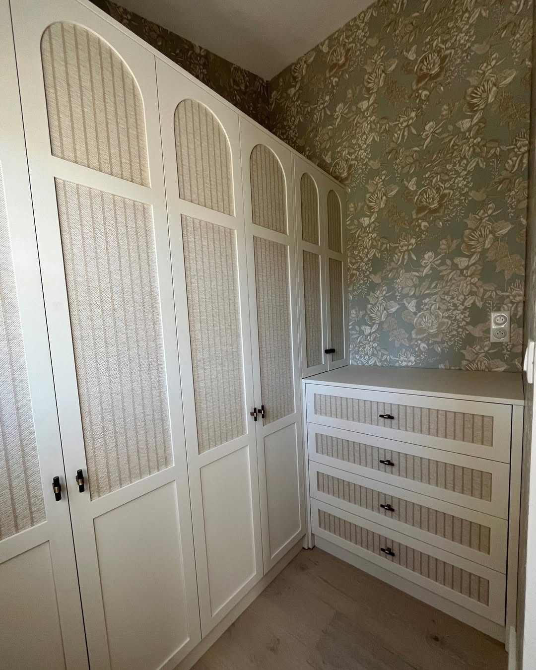 Rangement intégré blanc avec portes arquées à inserts texturés, jouxtant une commode assortie et un mur au papier peint floral vert sauge.