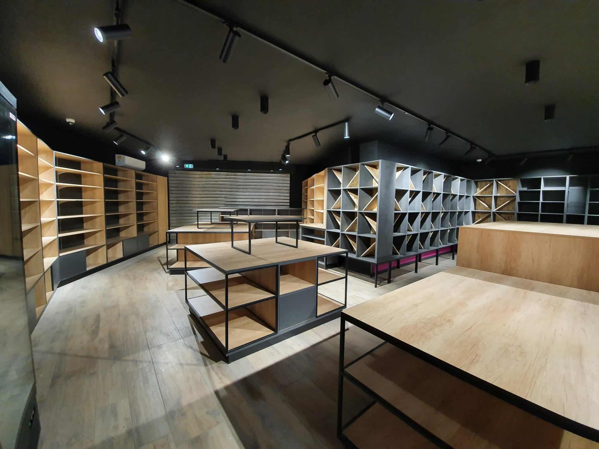 Intérieur d'un magasin ou entrepôt vide avec étagères en bois clair et métal noir, éclairage sur rail au plafond sombre. Le texte visible est "N".