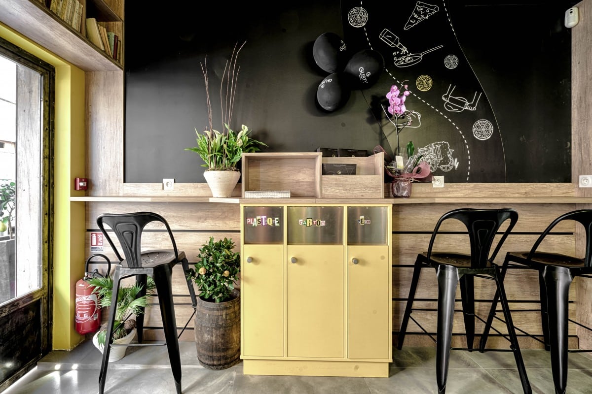 Coin recyclage jaune avec bacs "PLASTIQUE", "CARTON", "VERRE", chaises hautes noires, plantes, et tableau noir décoré.