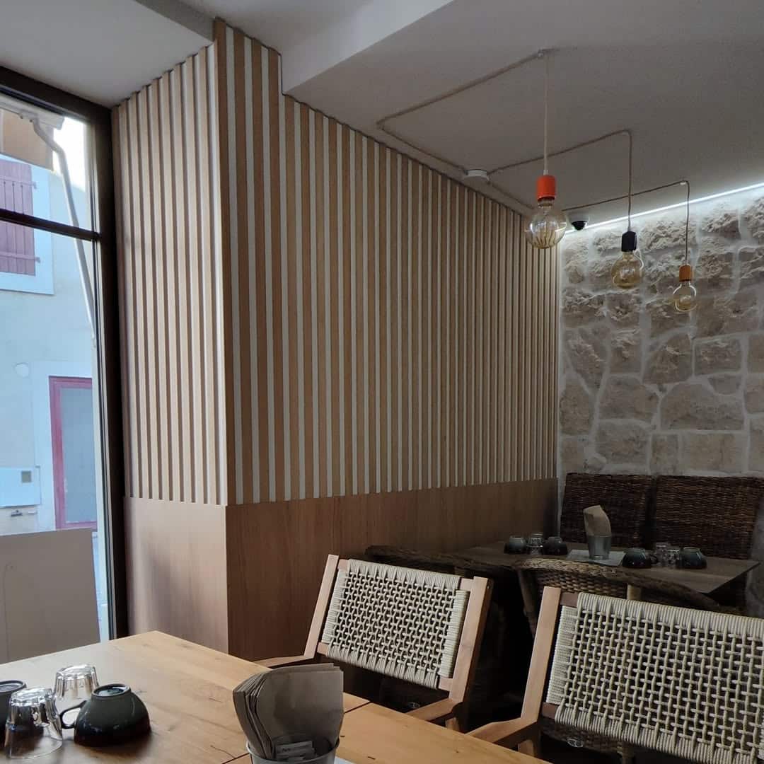Intérieur moderne de restaurant avec murs à lattes de bois clair, mur en pierre brute, chaises tressées, et suspensions lumineuses style industriel près d'une grande fenêtre.