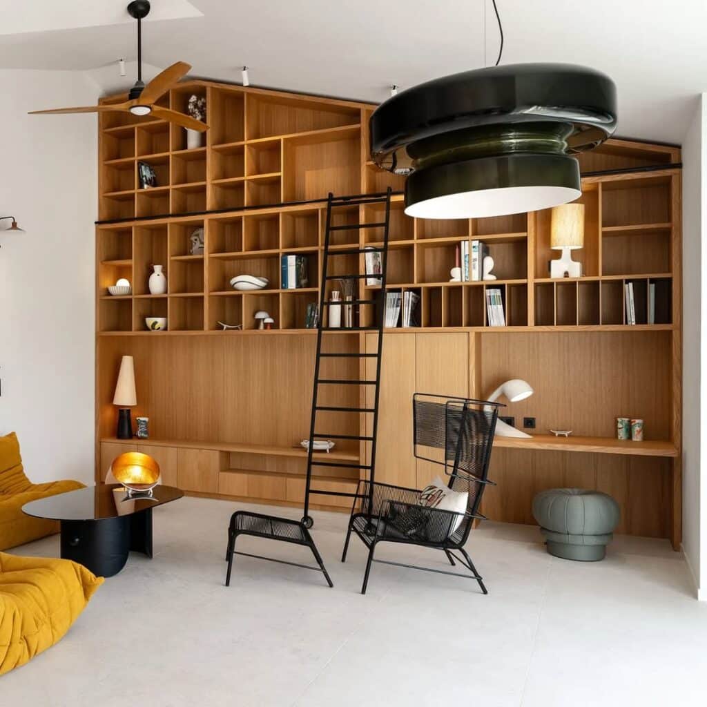 Bibliothèque bois et design moderne : salon cosy Intérieur moderne avec grande bibliothèque murale en bois clair, échelle roulante noire, fauteuils jaunes Togo, suspension noire, et sol clair.