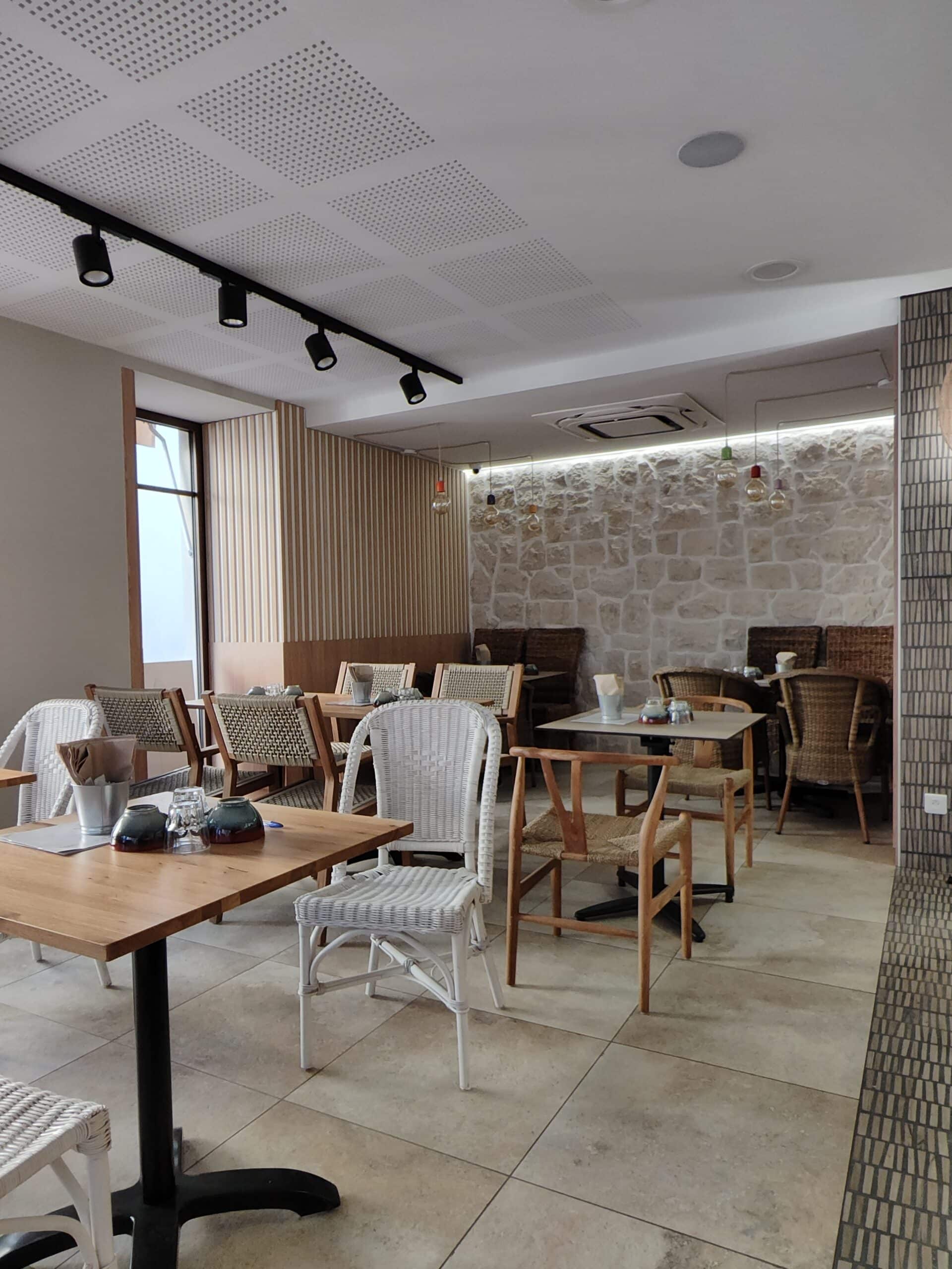 Salle de restaurant lumineuse avec tables en bois, chaises en osier blanc et mur rustique en pierres claires.