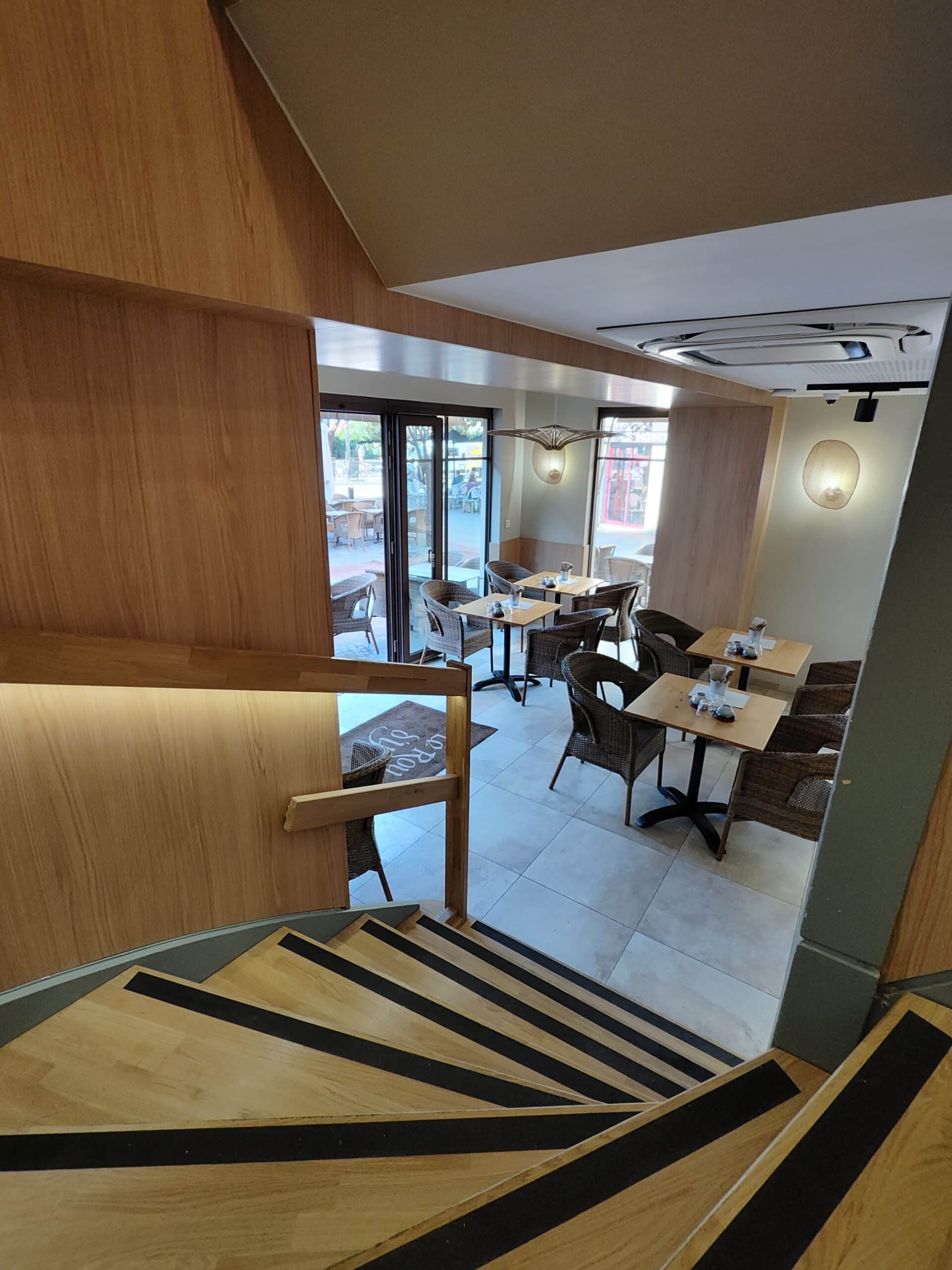Escalier en bois ouvrant sur la salle du restaurant Le Rou avec chaises en rotin et tables carrées modernes.