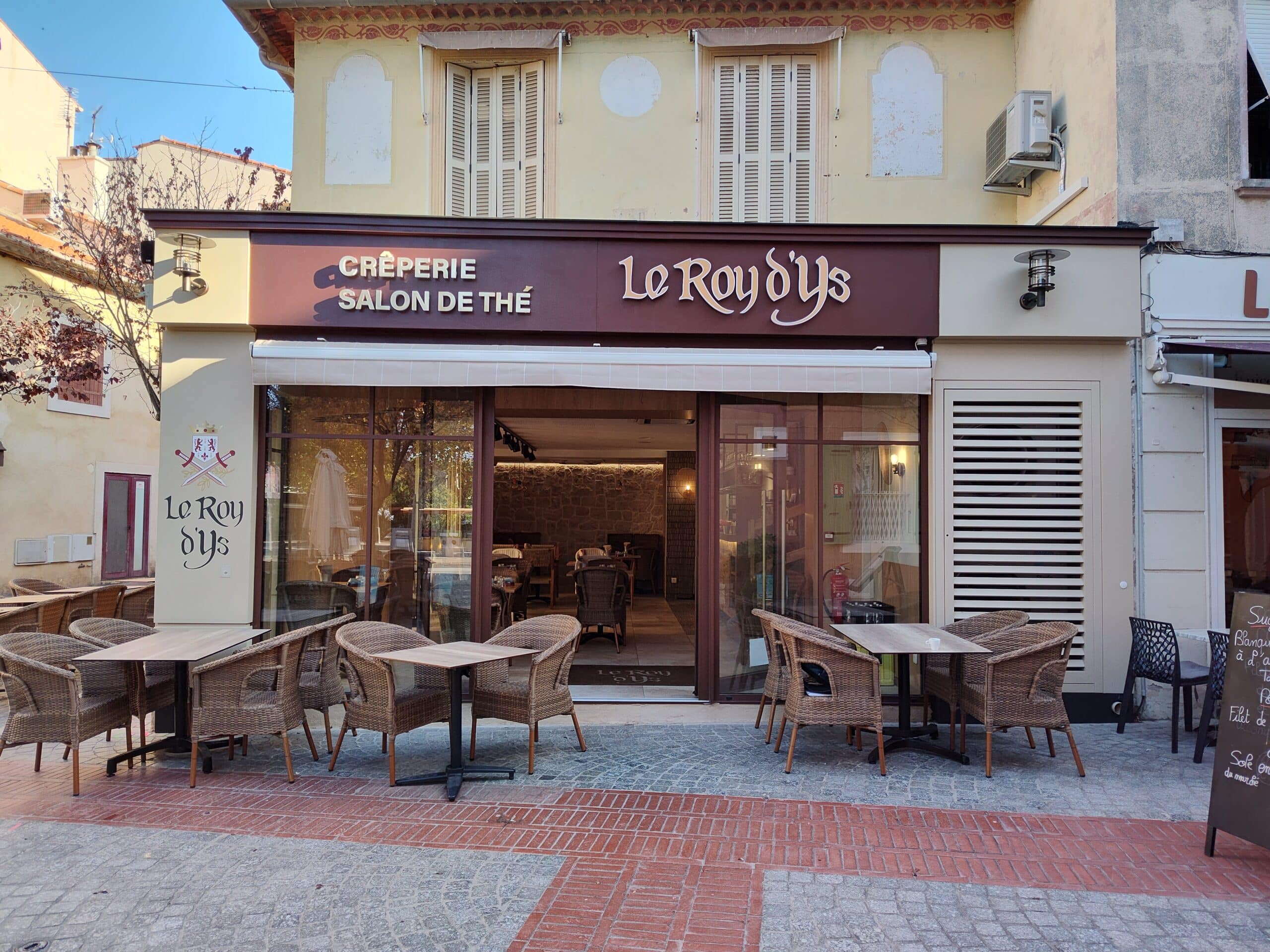 Façade de la crêperie salon de thé Le Roy d'Ys avec terrasse en osier et tables en bois sur rue pavée.