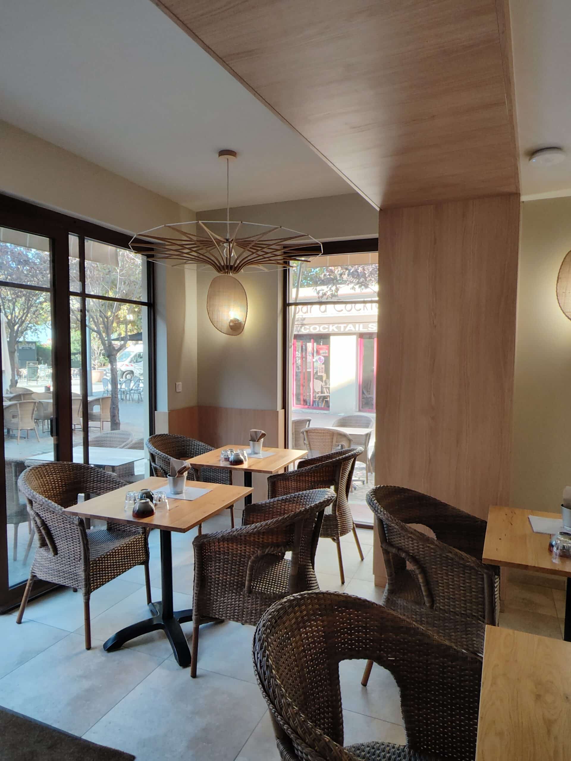 Restaurant lumineux avec chaises en rotin tressé, tables en bois massif et lustre moderne en bois près des fenêtres.