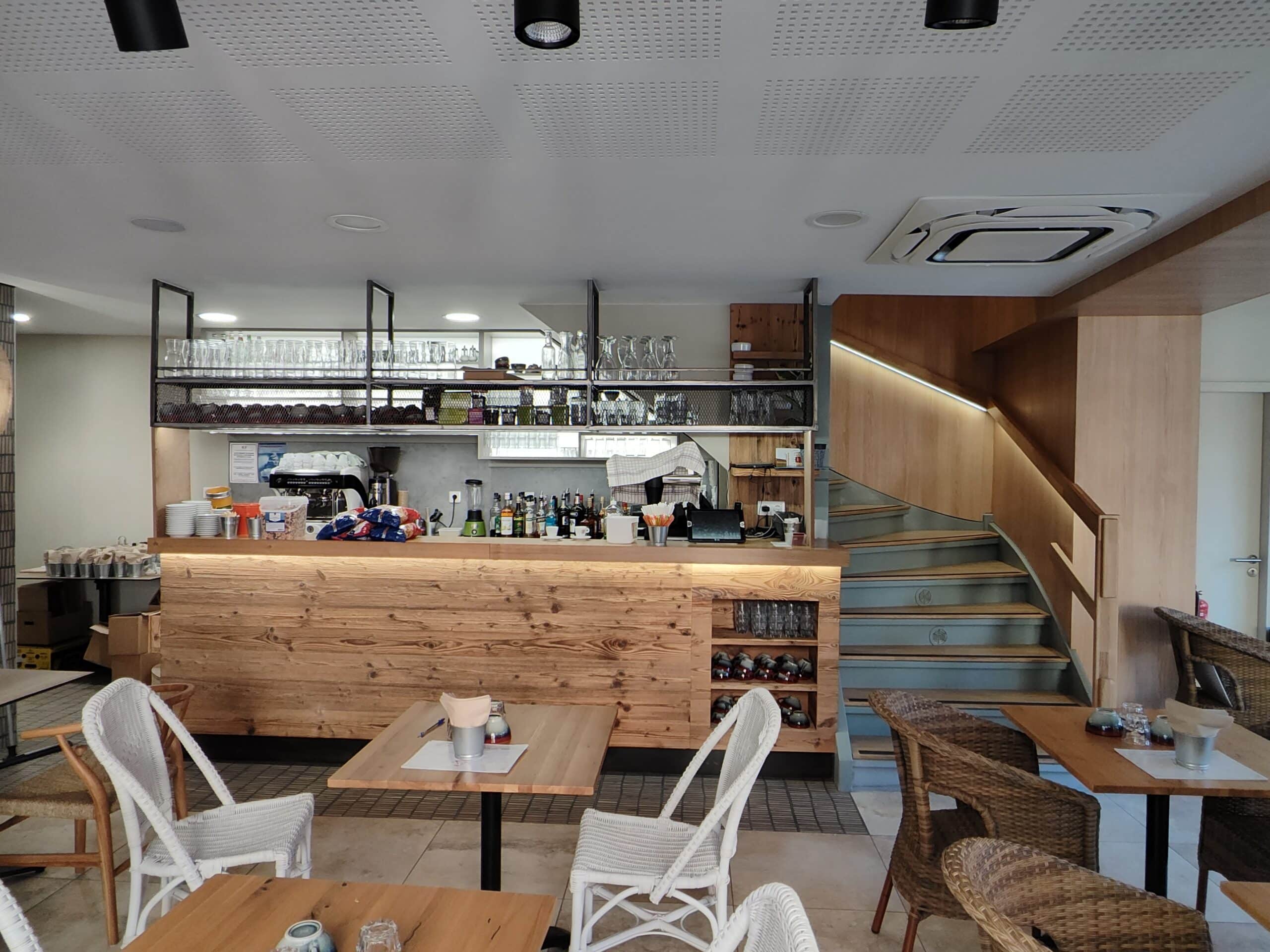 Intérieur de café moderne avec comptoir en bois massif, chaises en rotin et escalier design éclairé.