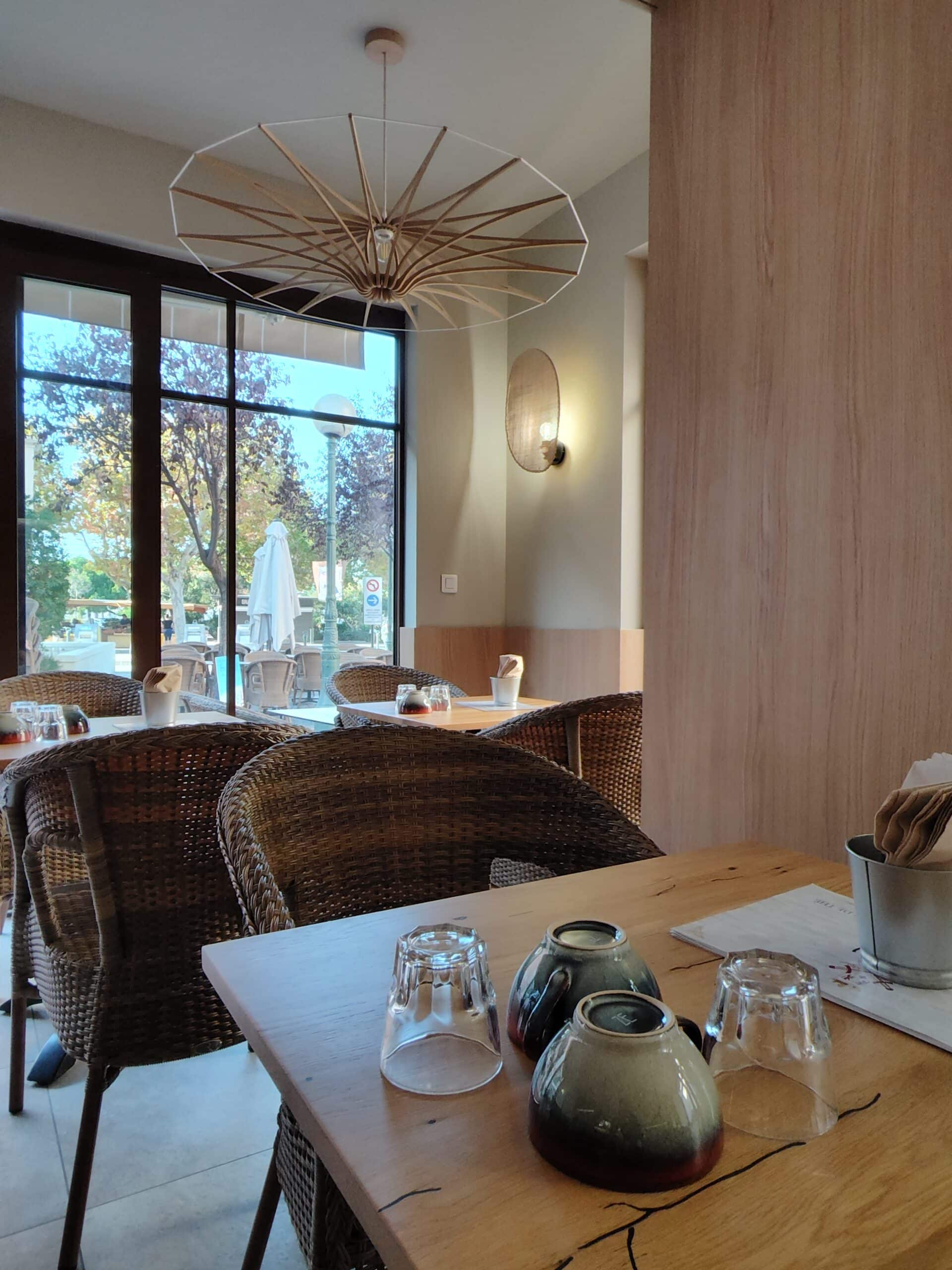Intérieur de restaurant chaleureux avec chaises en osier, tables en bois et grand luminaire design suspendu.