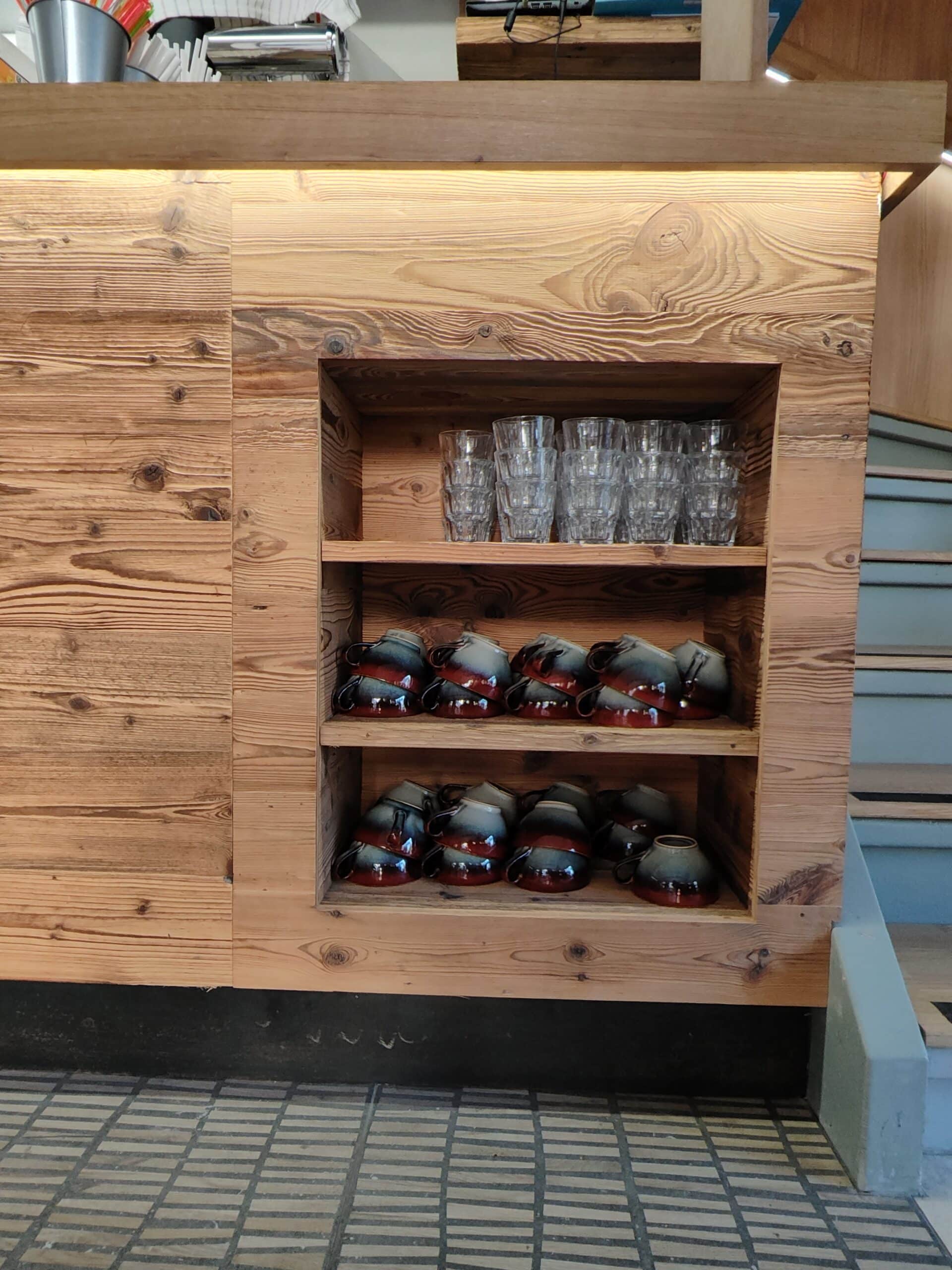 Étagère murale en bois avec tasses en céramique artisanale et verres dans un café au style rustique.